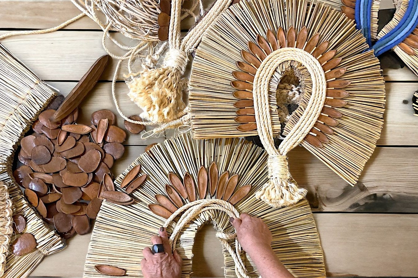 Mãos de uma artesã montando cocares de palha e sementes sobre a bancada do ateliê